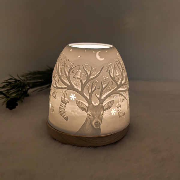 Beleuchtetes Lithophanie-Windlicht aus weißem Porzellan auf einem Holzsockel mit einem detailreichen Hirsch-Relief, dessen Geweih mit Glocken, Sternen und Socken verziert ist.